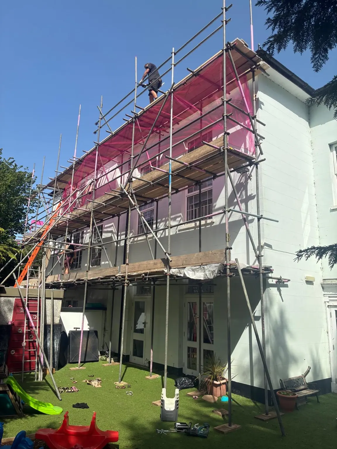 Multi-lift scaffold with pink debris netting above a nursery outdoor play area