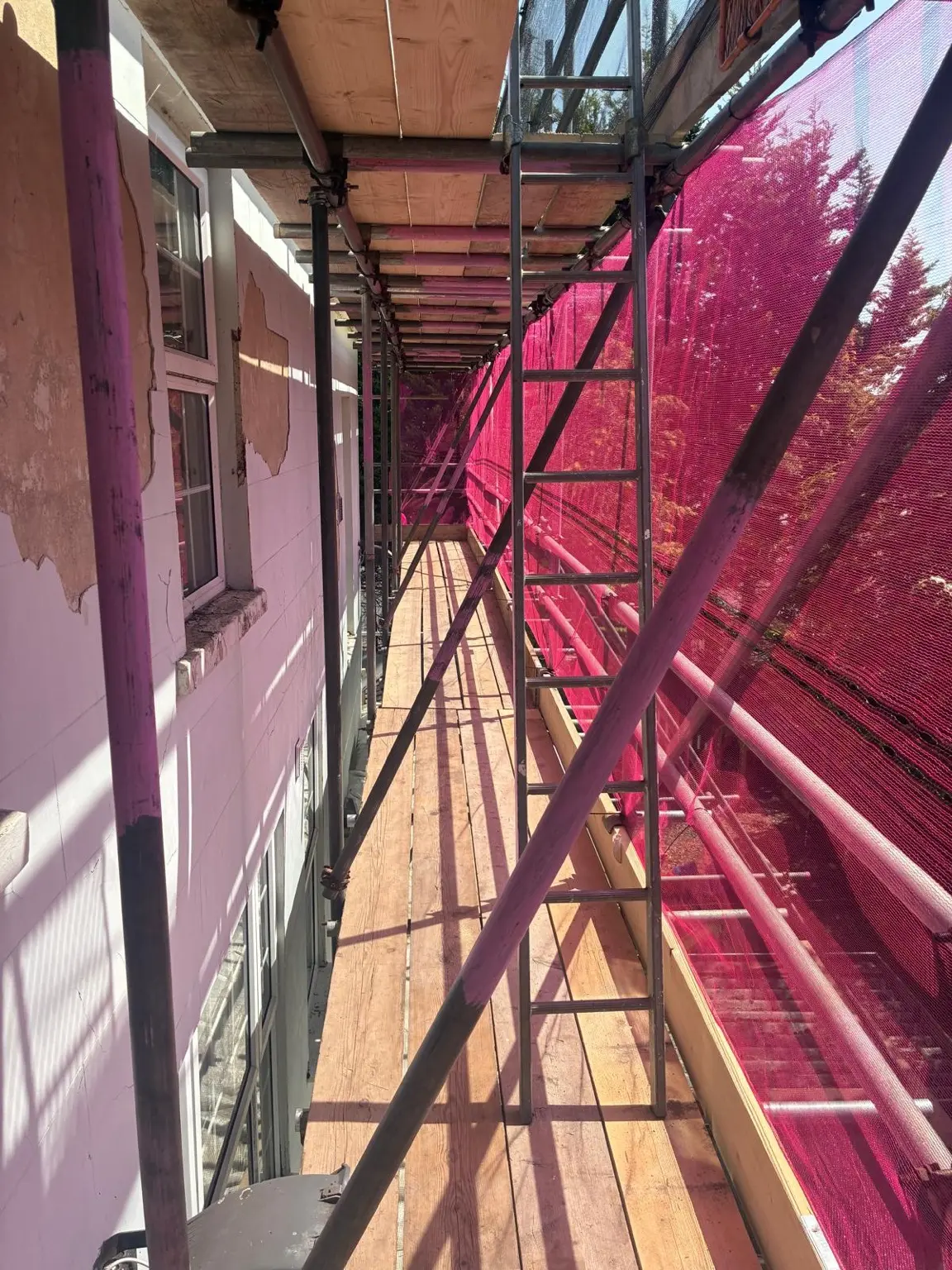 Boarded scaffold platform with guardrails and pink debris netting beside the nursery building